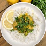 Tajine de poulet au citron et à la courgette servi avec du riz dans un bol blanc. Tranches de citron en garniture et citrons entiers et coriandre en arrière-plan.