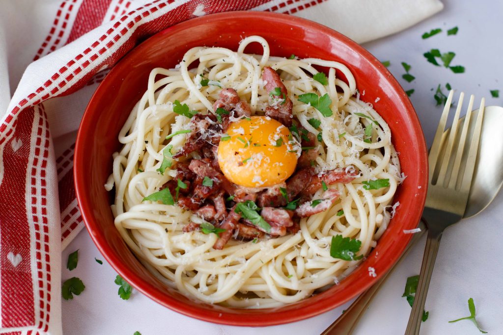 creamy pastat carbonara shown in a red bowl with lardons and an egg yolk on top