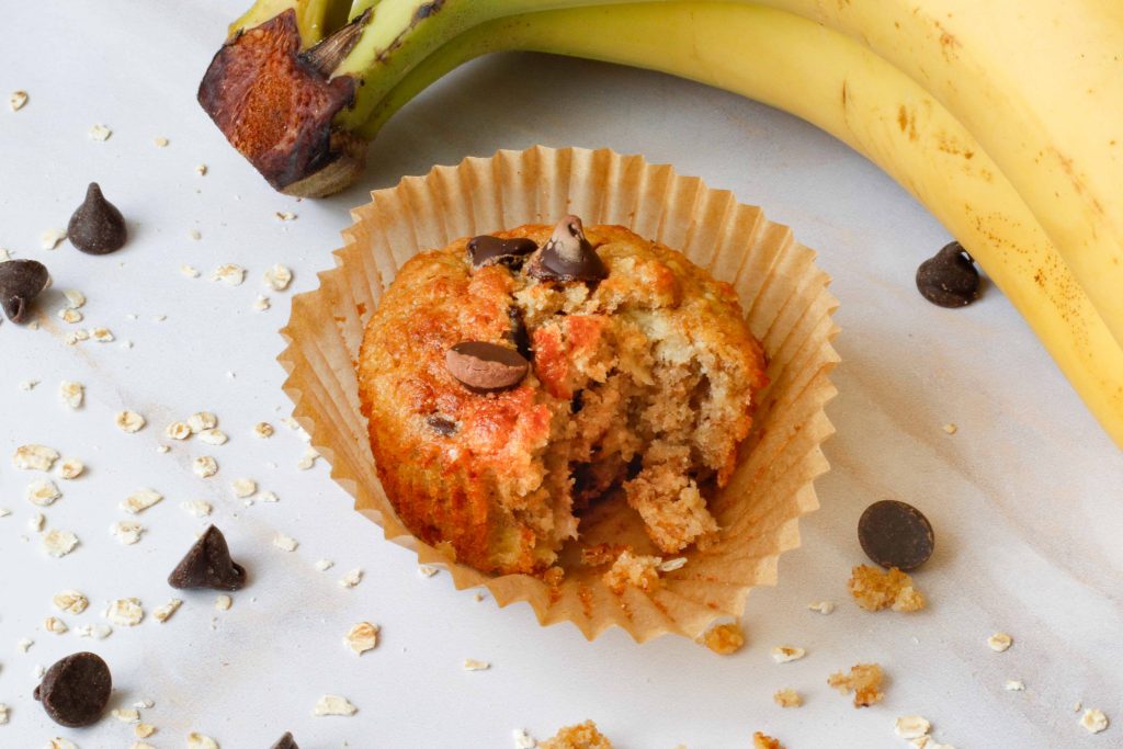 close up of Sourdough Banana Oat Muffins with Chocolate Chips bitten into with bananas and choco chips in background